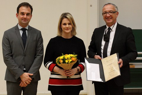 Drei Personen stehen nebeneinander, in der Mitte eine junge Frau. Sie hält Blumen in der Hand. Der Mann rechts neben ihr hält eine aufgeklappte Urkunde in der Hand.