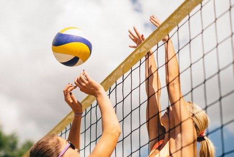 Zwei Frauen spielen Volleyball. Beide haben ihre Arme weit oben bzw. über dem Netz. Der Ball befindet sich in der Luft. Das Foto ist eine Nahaufnahme. Das Volleyballnetz geht vom linken unteren Bildrand bis zum rechten oberen Rand.
