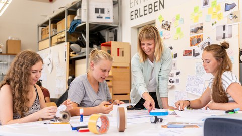 Das Farbfoto zeigt eine Personengruppe an einem großen Arbeitstisch. Drei Mädchen schauen auf den Tisch. Die vierte junge Frau steht lächelnd daneben und zeigt auf den Tisch.