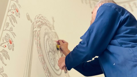 Das Farbfoto zeigt einen Restaurator im blauen Kittel, der gerade ein Motiv auf eine Wand malt.
