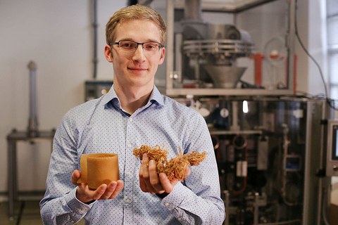 Das Foto zeigt den Nachwuchswissenschaftler Ludwig Schmidtchen, der in seinen Händen hellbraune, getrocknete Algen hält. Im Hintergrund ist eine Laborapparatur zu sehen.