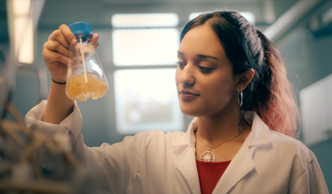Auf dem Bild ist eine Studierende zu sehen, die ein Reagenzglas mit Flüssigkeit in der Hand hält