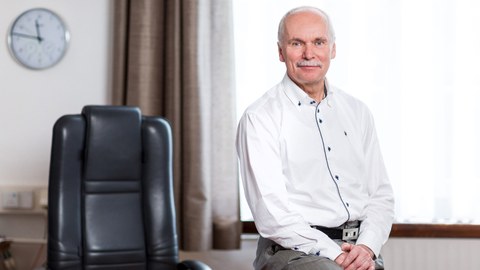Das Foto zeigt Prof. Ralph Stelzer, der seitlich auf seinem Dekane-Schreibtisch sitzt. Er trägt ein weißes Hemd und eine graue Hose.