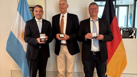 Drei Männer mittleren Alters in Anzügen posieren mit ihren Preisen. Im Hintergrund sind die argentinische und deutsche Flagge.