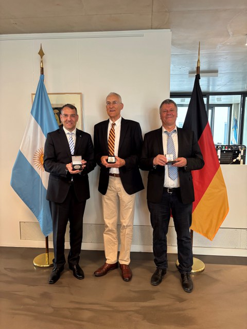 Drei Männer mittleren Alters in Anzügen posieren mit ihren Preisen. Im Hintergrund sind die argentinische und deutsche Flagge.