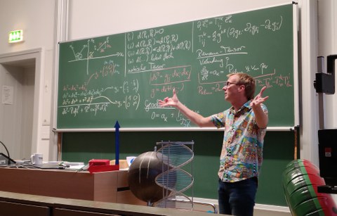 Eine Person steht vor einer mit mathematischen Formeln beschriebenen grünen Tafel.