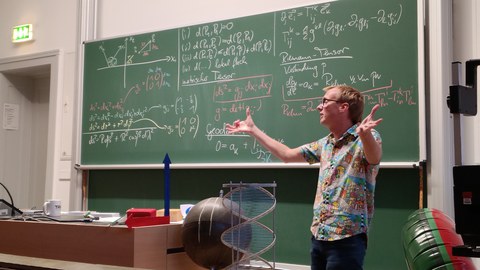 Eine Person steht vor einer mit mathematischen Formeln beschriebenen grünen Tafel.