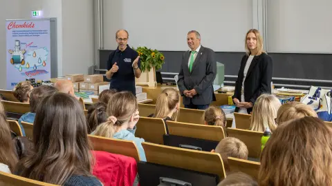 Kinder sitzen in einem Hörsaal und schauen Richtung Podium. Dort sind drei Personen zu sehen, die einen Vortrag halten.