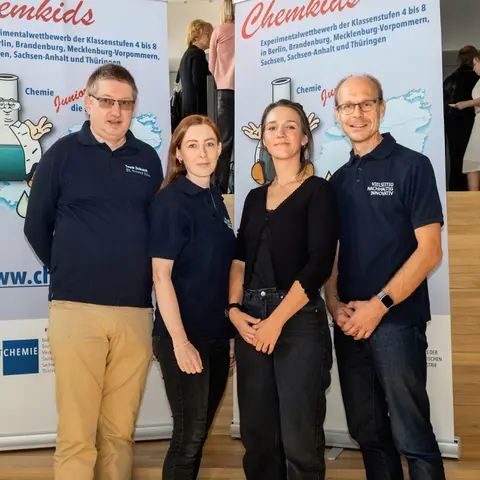 Eine Gruppe von vier Personen, die vor zwei Roll-Ups stehen, die den Hinweis auf den Experimentalwettbewerb ChemKids geben.