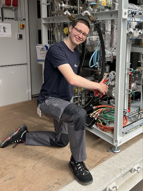 Junger Mann mit kurzen Haaren und Brille in Arbeitskleidung hantiert mit einer Zange an einer Mutter in einer Werkstatt. Er lächelt in die Kamera.