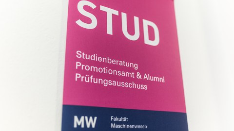 Foto:  Hinweisschild mit STUD (in sehr großer Schrift), darunter steht Studienberatung, darunter Promotionsamt & Alumni und darunter Prüfungsausschuss, am unteren Rand des Schildes steht MW Fakultät Maschinenwesen