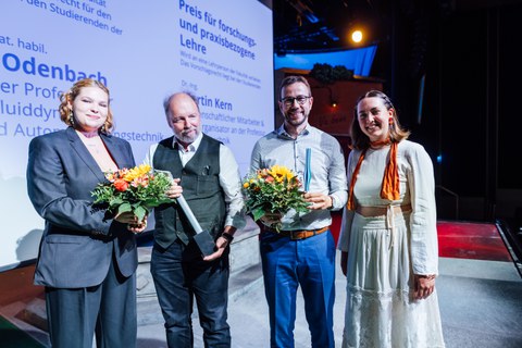 Zwei Männer mit Blumensträußen und Pokalen flankiert von zwei studentischen Vertreterinnen des Fachschaftsrats 