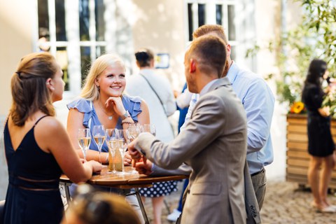 Gruppe von Absolventen und Absolventinnen unterhält sich - zentriert eine junge Frau mit blonden Haaren in blauem Kleid