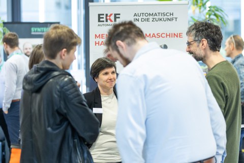 Mehrere Personen lassen sich von Frau mit dunklem Haar und schwarmen Blazer an einem Stand von EKF beraten