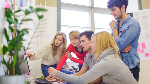 Foto: In einem Arbeitsraum sitzen an einem Schreibtisch 4 junge Personen und ein junger Mann steht hinter ihnen. Sie diskutieren miteinander und zeigen dabei auf etwas am Bildschirm was im Foto aber nicht zu sehen ist