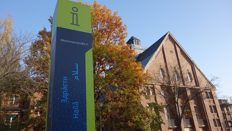 Das Foto zeigt eine Gebäude und eine Informationssäule an der Mommsenstraße 9. Auf der Informationstafel ist das Wort "Hallo" in verschiedenen Sprachen geschrieben. Die Blätter des Baumes im Hintergrund sind bunt gefärbt. 
