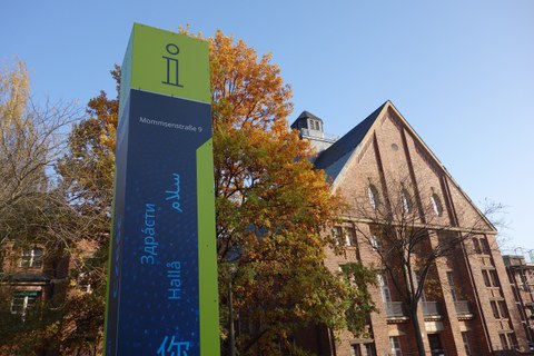 Das Foto zeigt eine Gebäude und eine Informationssäule an der Mommsenstraße 9. Auf der Informationstafel ist das Wort "Hallo" in verschiedenen Sprachen geschrieben. Die Blätter des Baumes im Hintergrund sind bunt gefärbt. 