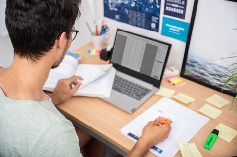 Das Foto zeigt einen Studenten an seinem Schreibtisch. Vor ihm steht sein Laptop und viele weitere Büromaterialien. Mit einem Stift notiert er gerade etwas auf einem Block. 