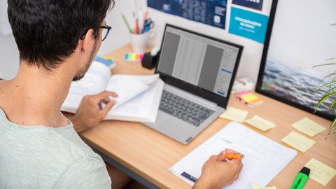 Das Foto zeigt einen Studenten an seinem Schreibtisch. Vor ihm steht sein Laptop und viele weitere Büromaterialien. Mit einem Stift notiert er gerade etwas auf einem Block. 
