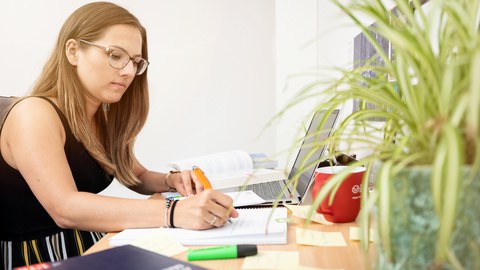  Das Foto zeigt eine Studentin an ihrem Schreibtisch. Vor ihr steht ihr Laptop und viele weitere Büromaterialien. Mit einem Stift notiert sie gerade etwas auf einem Block.