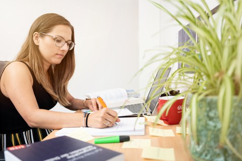  Das Foto zeigt eine Studentin an ihrem Schreibtisch. Vor ihr steht ihr Laptop und viele weitere Büromaterialien. Mit einem Stift notiert sie gerade etwas auf einem Block.