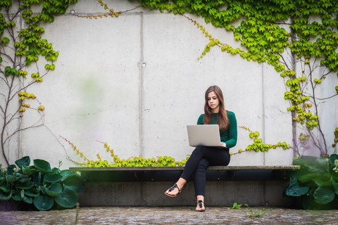 Das Bild zeigt eine Studentin mit einem Notebook. Sie sitzt auf einer Bank vor einer mit Grünpflanzen bewachsenen Wand.