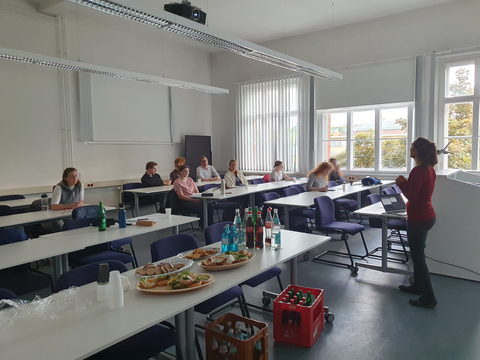 Menschen die an Tischen sitzen, während eine Referentin spricht. Im Vordergrund Getränke und belegte Brötchen
