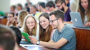 Das Foto zeigt Studierende im Hörsaal. Im MIttelpunkt des Betrachters stehen drei Studierende, die gemeinsam auf ein Tablett schauen.