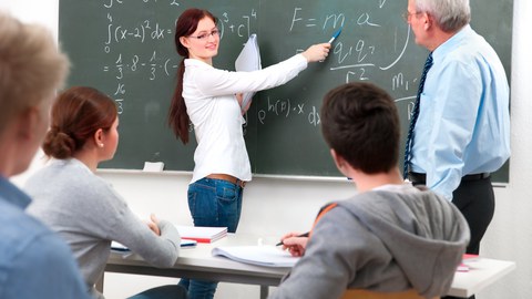 Auf dem Bild sieht man eine Gruppe von Studierenden mit ihrem Dozenten. Eine Studierende erklärt an der Tafel die Herleitung einer Formel.