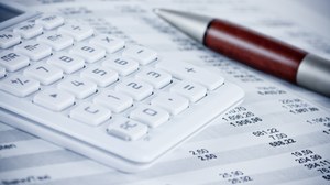 Auf dem Foto sieht man einen weißen Taschenrechner und einen Kugelschreiber auf einem Blatt Papier mit Tabellenkalkulationen liegen.