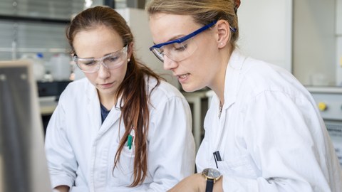 Foto von zwei Frauen im Labor. Beide tragen Arbeitskleidung und Schutzbrillen