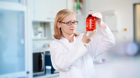 Junge Wissenschaftlerin mit Brille hält eine Flasche mit roter Flüssigkeit in den Händen und betrachtet sie. 