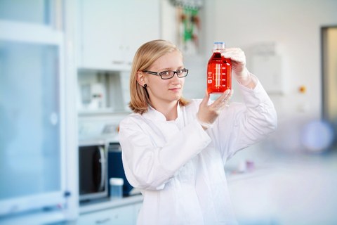 Junge Wissenschaftlerin mit Brille hält eine Flasche mit roter Flüssigkeit in den Händen und betrachtet sie. 