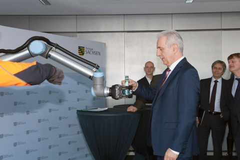 Auf dem Foto sieht man den ehemaligen Ministerpräsidenten Stanislaw Tillich während der Pressekonferenz zum Smart Systems Hub Dresden, wie er von einem Roboterarm eine Flasche Wasser entgegennimmt.