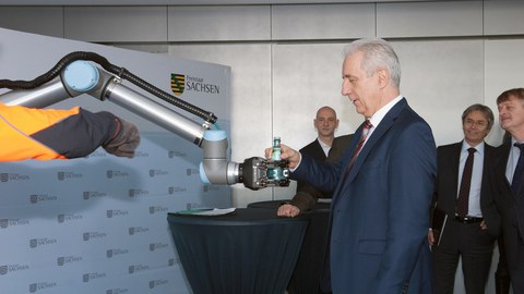 Auf dem Foto sieht man den ehemaligen Ministerpräsidenten Stanislaw Tillich während der Pressekonferenz zum Smart Systems Hub Dresden, wie er von einem Roboterarm eine Flasche Wasser entgegennimmt.