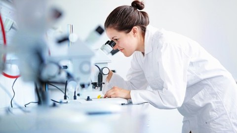 Auf dem Foto sieht man eine junge Wissenschaftlerin im weißen Kittel, die durch ein Mikroskop schaut.