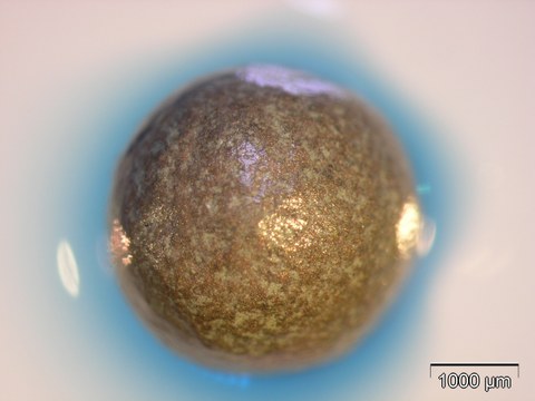 Auf dem Foto sieht man eine goldfarbene Kugel, die von einem blauen Schein (Corona) umgeben ist. Es handelt sich dabei um eine metallische Hohlkugel mit Pilzenzymen. Die Enzymreaktion (blaue Corona) ist mit einem Farbstoff sichtbar gemacht.