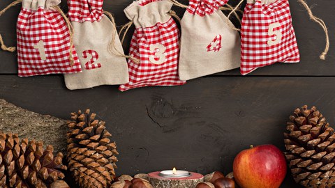 Auf dem Bild sieht man in der oberen Hälfte fünf Jute-Säckchen eines Adventskalenders. Darunter sind Tannenzapfen, Haselnüsse, ein Teelicht und ein Apfel abgebildet.