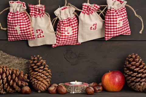Auf dem Bild sieht man in der oberen Hälfte fünf Jute-Säckchen eines Adventskalenders. Darunter sind Tannenzapfen, Haselnüsse, ein Teelicht und ein Apfel abgebildet.