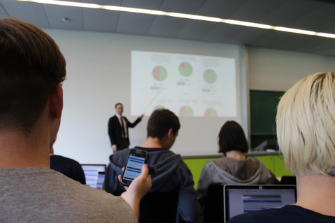 Man sieht Personen in einem Seminarraum, die Richtung Podium schauen. Dort erklärt eine Person eine Präsentation.