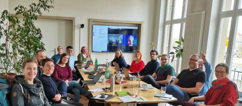 Auf dem Foto sind 14 Menschen abgebildet, die an einem Tisch gemeinsam arbeiten. Im Hintergrund sieht man eine Person digital zugeschaltet.