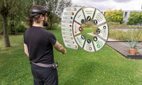 The picture shows a collage of photo and 3D visualization. A man stands on a meadow with 3D glasses and looks virtually at a special user interface. 