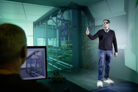Ein junger Mann steht in einen Raum zur Projektion einer dreidimensionalen Illusionswelt der virtuellen Realität. Er hat eine Datenbrille auf und einen Controler in der Hand. Im Vordergrund sitzt ein Mitarbeiter vor einem Computer.
