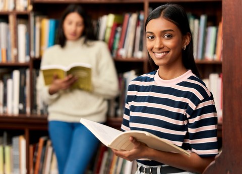 Foto von zwei Studentinnen in der Bibliothek. Die eine im Hintergrund liest und die im Vordergrund lächelt in die Kamera. 