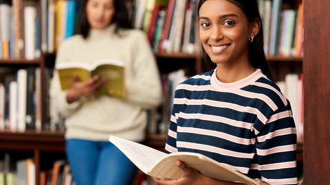 Foto von zwei Studentinnen in der Bibliothek. Die eine im Hintergrund liest und die im Vordergrund lächelt in die Kamera. 