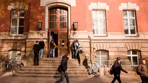Auf dem Foto sieht man den Haupteingang des Zeuner-Baus. Studierende verlassen das Gebäude. 