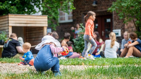 Foto eines Kleinkindes, das mit dem Rücken zum Betrachter auf einer Wiese hockt und einen Greifball hält. Im Hintergrund sind weitere Kinder mit ihren Eltern, Gebäude, Bäume und Wiese zu sehen.