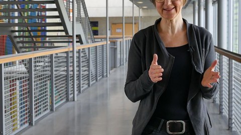  The picture shows Dr. Sandra Buchmüller in the main lecture hall. In the background, you can see a stairway and a colorful wall painting.