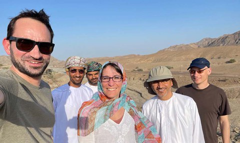 A group of six people standing together in a desert landscape with mountains in the background. They are smiling at the camera; some are wearing sunglasses, traditional clothing, and hats. The sky is clear and blue.