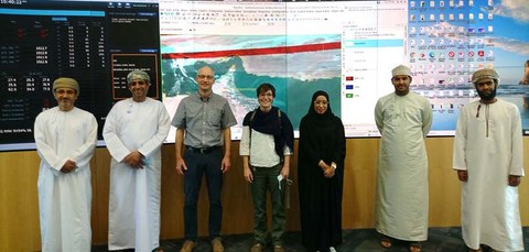 A group of seven people, wearing a mix of traditional Omani attire and casual Western clothing, stand in front of large display screens showing geographic data and computer desktops. They are smiling and standing in a well-lit room, likely a tech or data center.